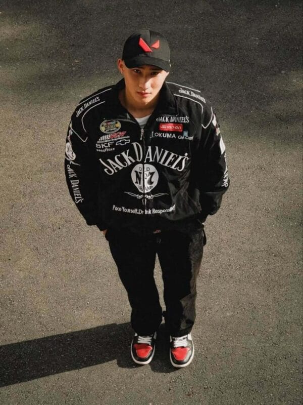 Man wearing a black Jack Daniel's racing jacket with red and black sneakers and a black cap with a red logo. He stands on a street surface looking up at the camera.