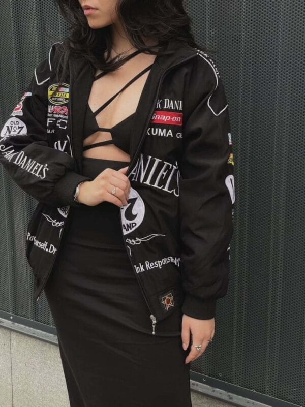Woman wearing a black Jack Daniel's racing jacket over a strappy black top and dress. Jacket features multiple sponsor patches and white embroidered text. She stands against a perforated metal wall.