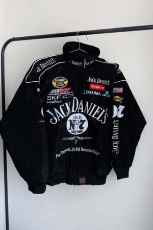 Front view of a black Jack Daniels jacket hanging on a rack. Features bold branding, logos of sponsors like Snap-on and Goodyear, and decorative racing patches on the sleeves.