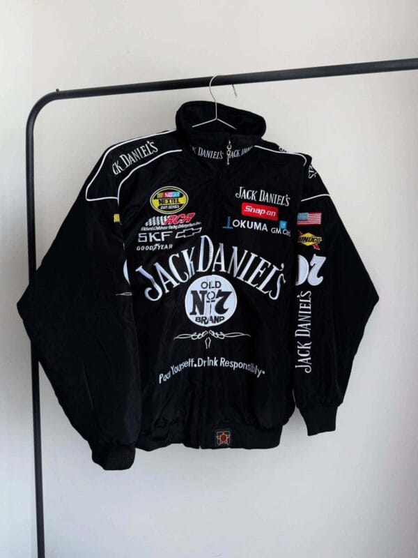 Front view of a black Jack Daniels jacket hanging on a rack. Features bold branding, logos of sponsors like Snap-on and Goodyear, and decorative racing patches on the sleeves.