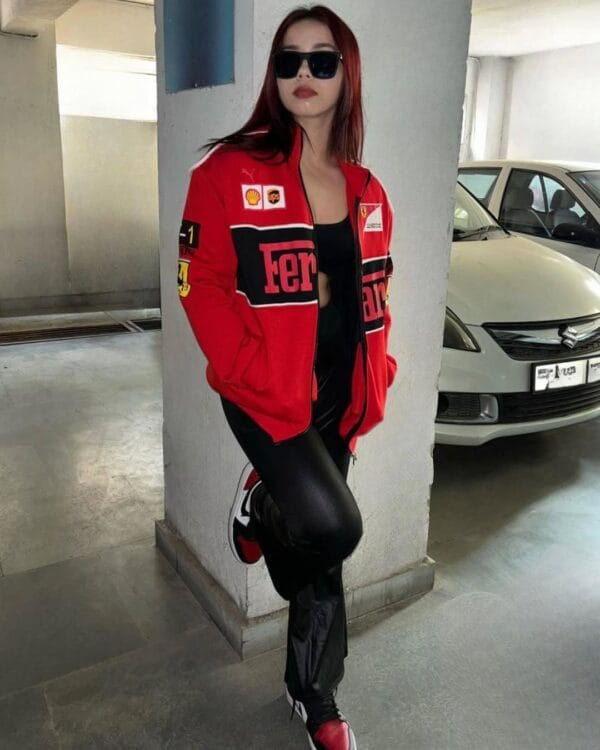 Woman posing in parking area wearing red Ferrari racing jacket, black leather pants, and sneakers, representing premium F1 apparel by Exotix.