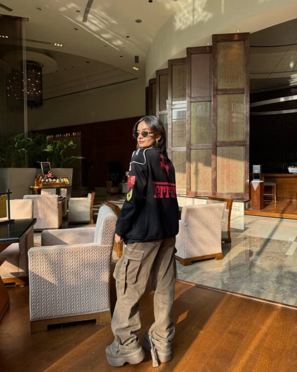 Woman wearing black Ferrari jacket by Exotix in a luxury hotel lounge with sunlight casting soft shadows – Formula 1 racing streetwear