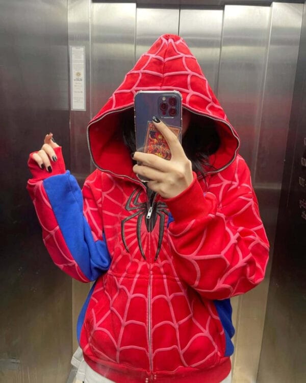 A young woman with long hair takes a mirror selfie while wearing a red and blue Spiderman-patterned hoodie in an elevator.