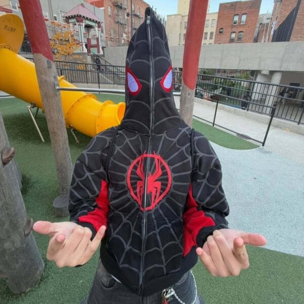 A young man wearing a red Spiderman-inspired hoodie with a grey stripe and red spider design, adjusting the collar while standing on a city street near a sticker-covered pole.