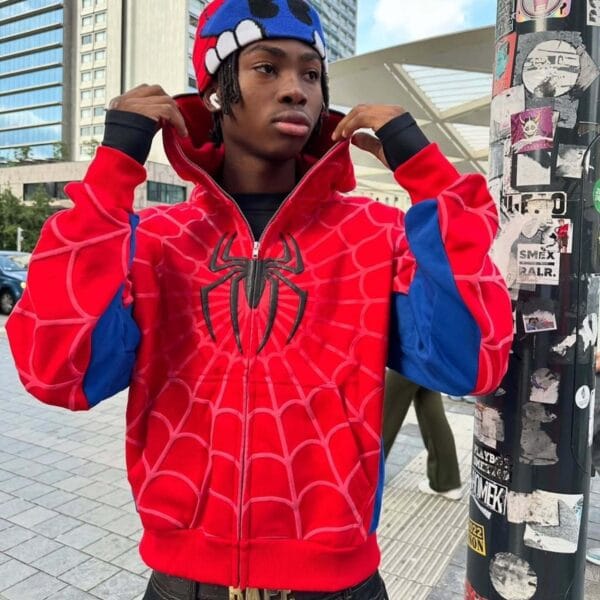 A man in a red Spidey hoodie posing with his hands on the hoodie’s collar, standing on a tiled pavement with a city building and blue sky in the background.