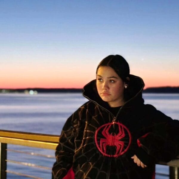 A woman with long hair wears a Spidey hoodie and takes a mirror selfie, showing off the hoodie’s front spider logo and vivid web pattern.