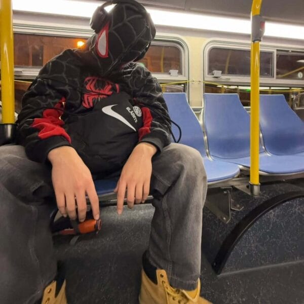 man leans against a concrete wall wearing a black Spidey hoodie with hands in his pockets, showcasing the casual streetwear style.