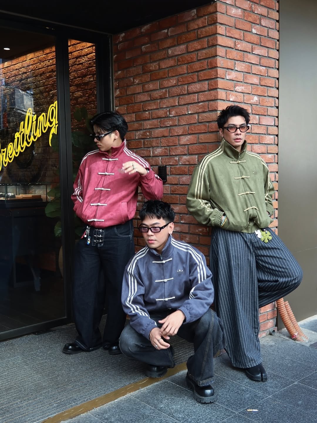Chinese New Year Jacket – Exotix CNY Edition Three models wearing Chinese New Year Jacket by Exotix in maroon, olive green, and dark grey, styled with striped pants and sunglasses against a brick wall storefront.