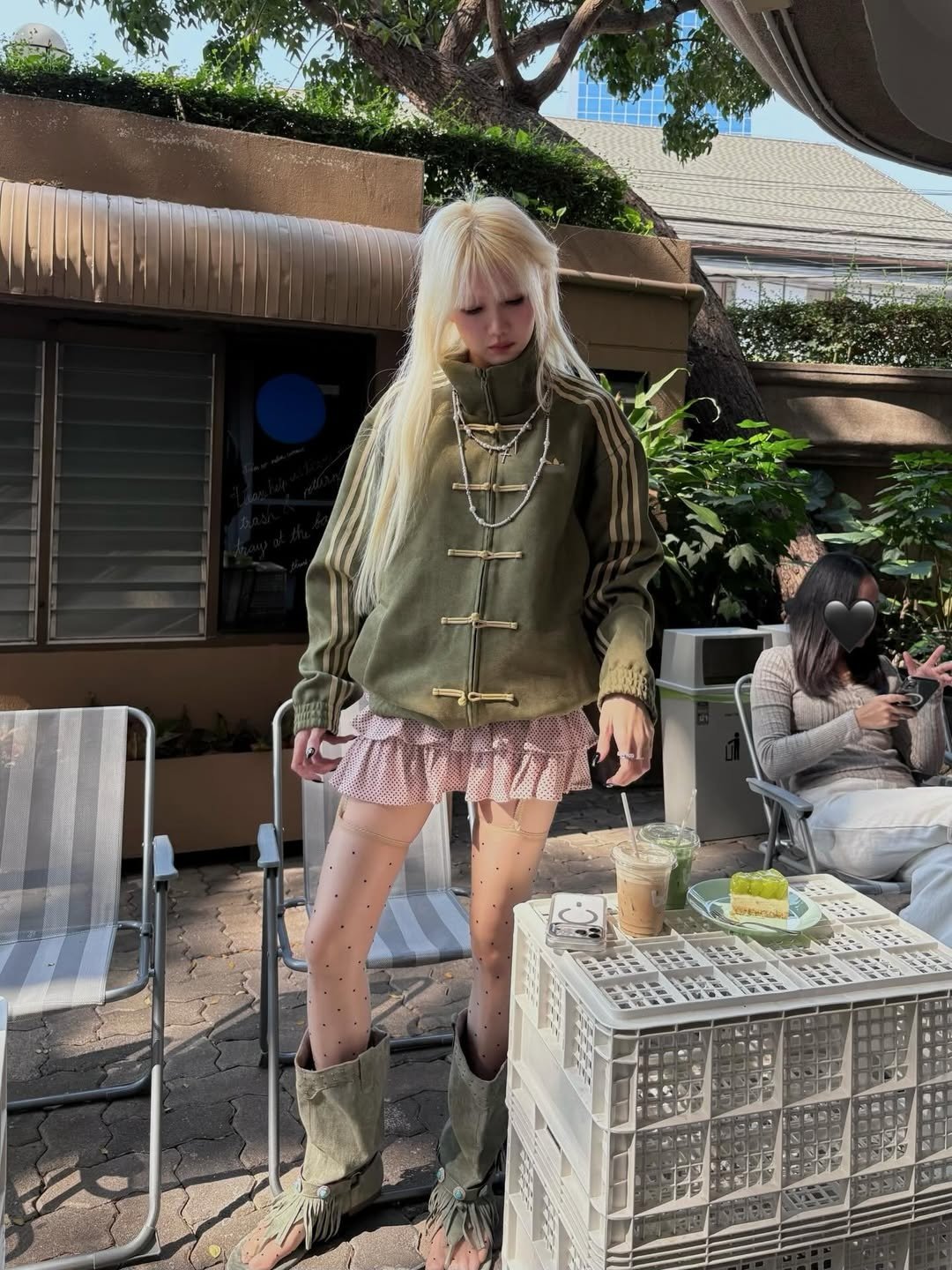 Chinese New Year Jacket – Exotix CNY Edition Woman styling an olive Chinese New Year jacket from exotix with a ruffled mini skirt, patterned tights, and fringe boots at an outdoor café.