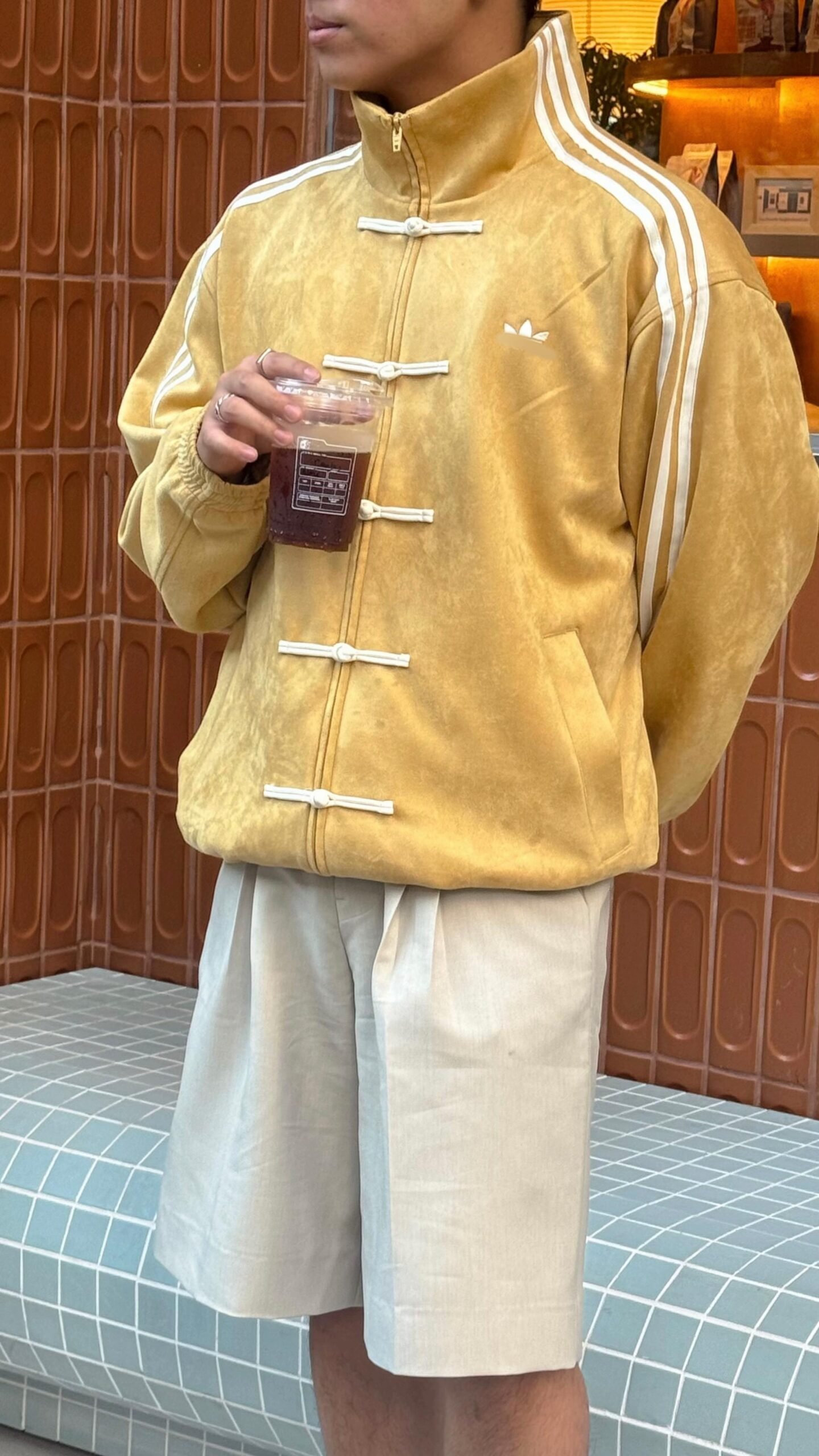 Chinese New Year Jacket – Exotix CNY Edition Close-up of a mustard yellow Chinese New Year jacket by exotix featuring traditional frog button details and striped sleeves, paired with beige shorts.