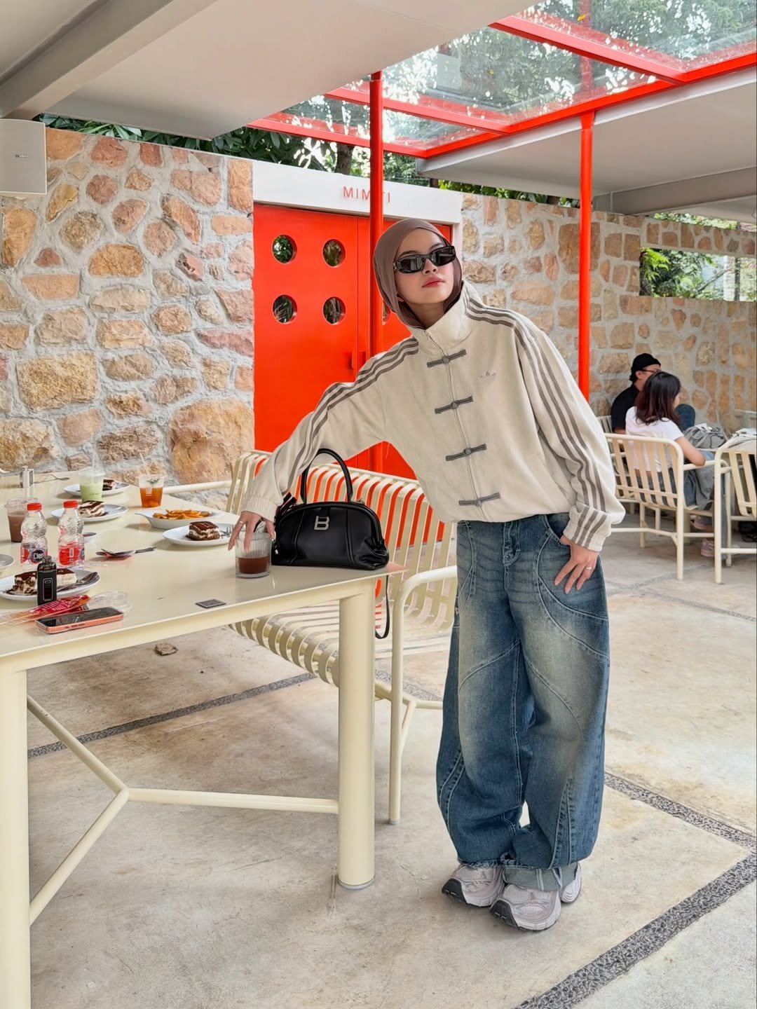 Chinese New Year Jacket – Exotix CNY Edition Woman wearing Exotix Chinese New Year jacket in beige with frog button details and sporty stripes, styled with wide-leg jeans and sunglasses at a modern café.